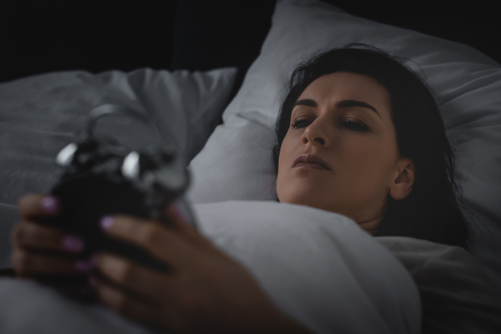 A woman lies in bed looking at the alarm clock she holds in her hands.